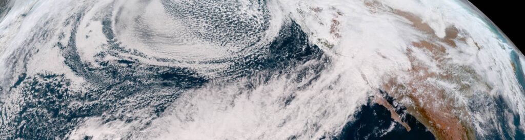 A satellite captures an atmospheric river from space. The white, banding plumes stretch across the Pacific and make landfall on the U.S. West Coast, with the curvature of Earth still in frame.