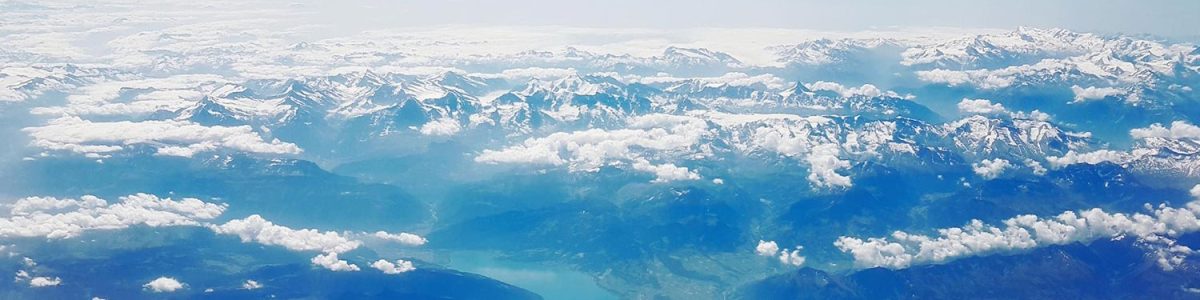 Aerial view of a mountain range with snow-capped peaks, clouds, and a lake nestled among the mountains.