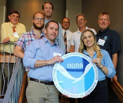 Delegation of eight NOAA employees standing on a staircase holding a large plaque with the NOAA emblem in front of them at the Eighth Biennial Southwest Aviation Weather Safety Workshop in Tempe, Arizona